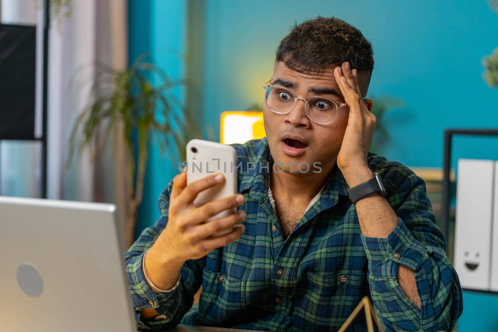 Sad Indian man use smartphone typing browsing, loses becoming surprised sudden lottery online game results, bad news, fortune loss, fail. Hispanic guy sitting at home office desk workplace. Lifestyle