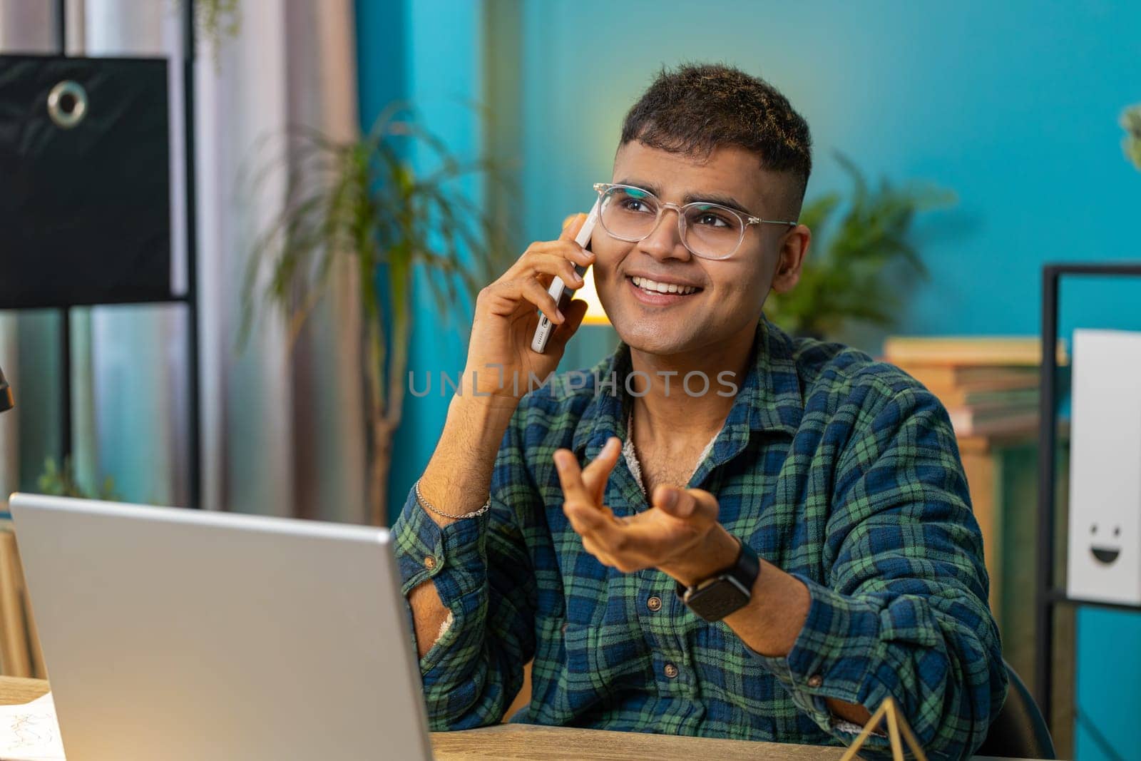 Indian man making phone call conversation with client or colleague friend. Arabian guy freelancer enjoying mobile loudspeaker talking gossip rumors, good news at home office table workplace. Lifestyle