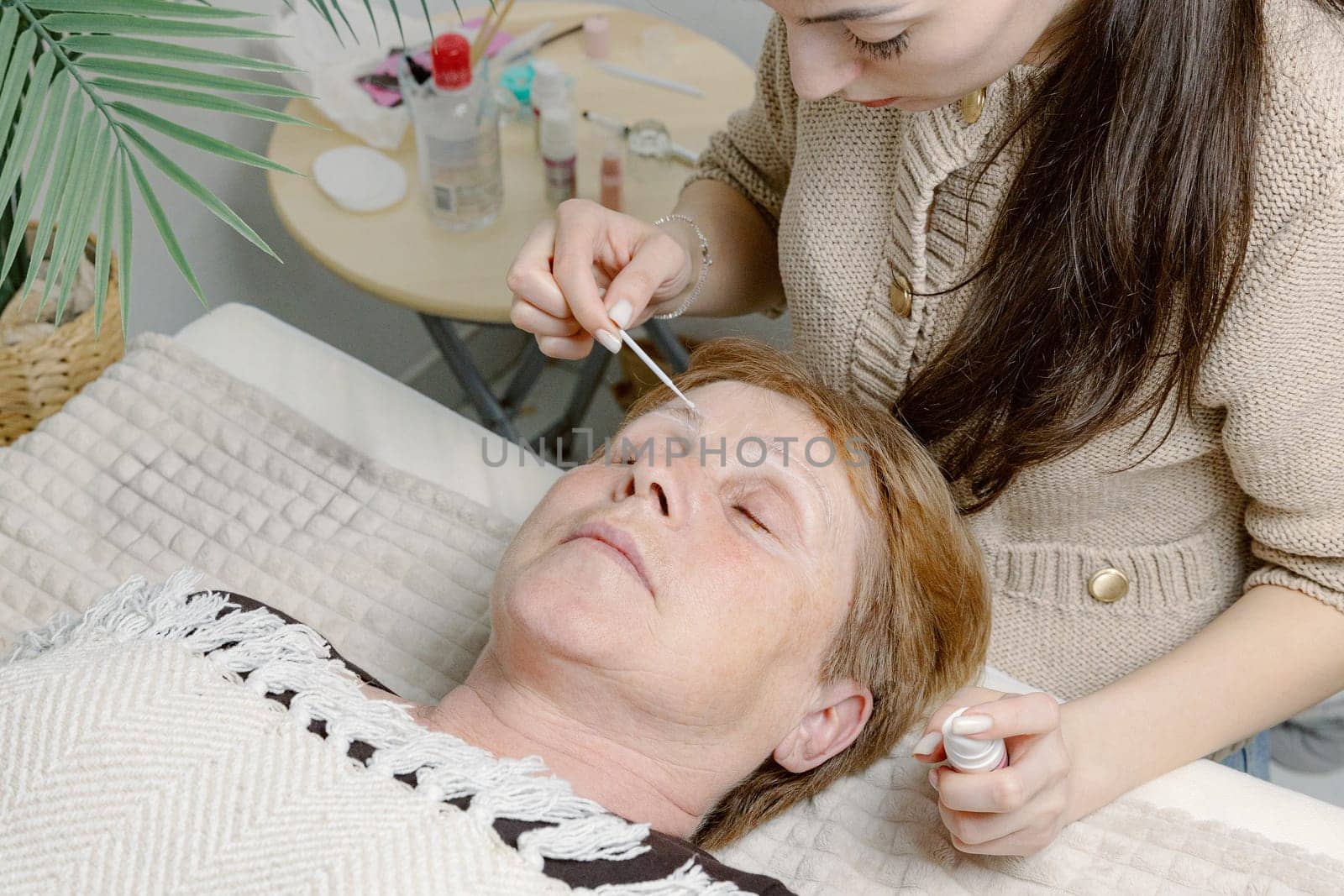 One young Caucasian young girl brunette cosmetologist degreases the eyebrows of an elderly woman with gray-blue eyes using a stick lying on a cosmetology bed in a home beauty salon, close-up side view.