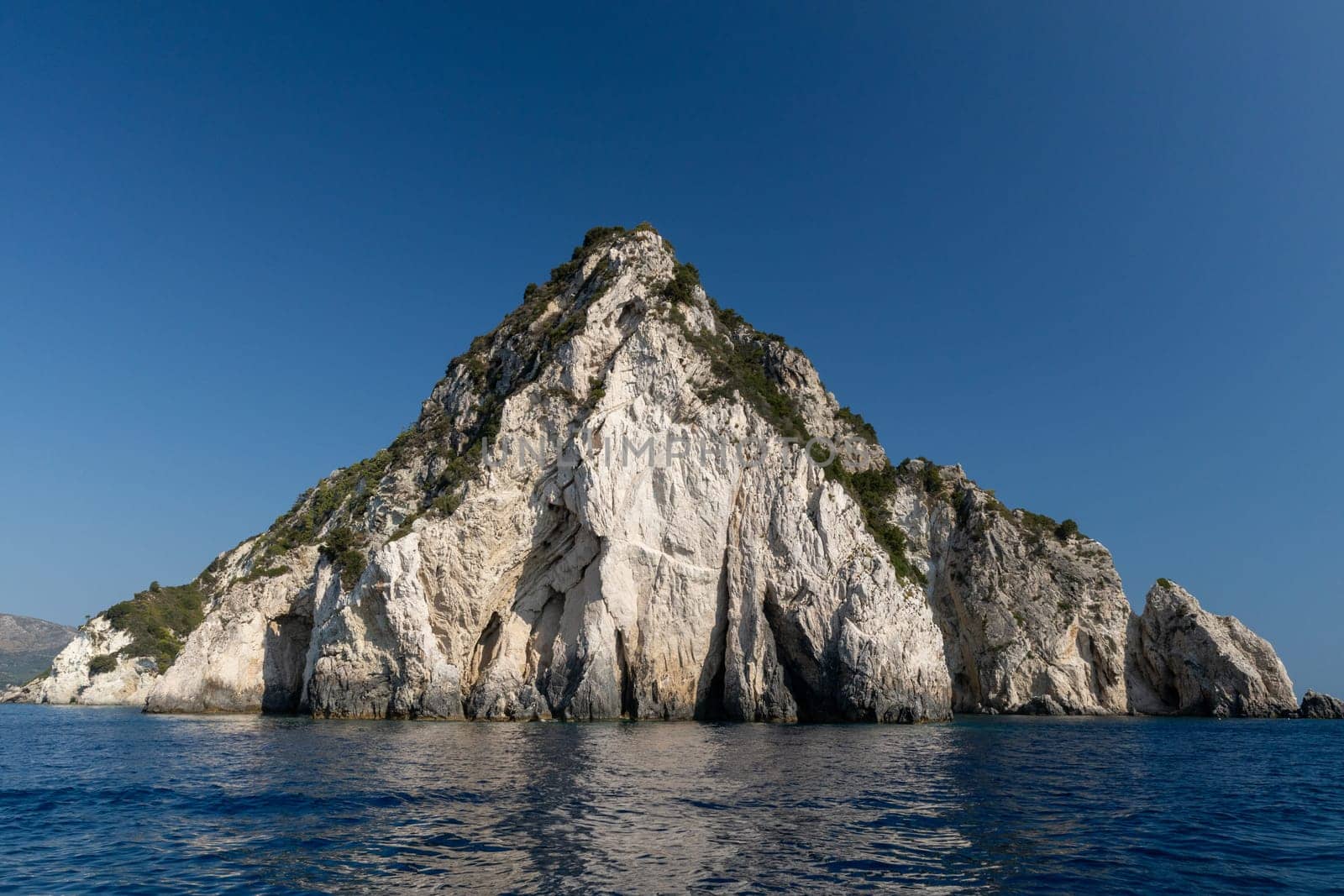 Beautiful panoramic view of a green rocky triangular cliff in the blue sea with a deserted beach on a sunny summer day, close-up side view.