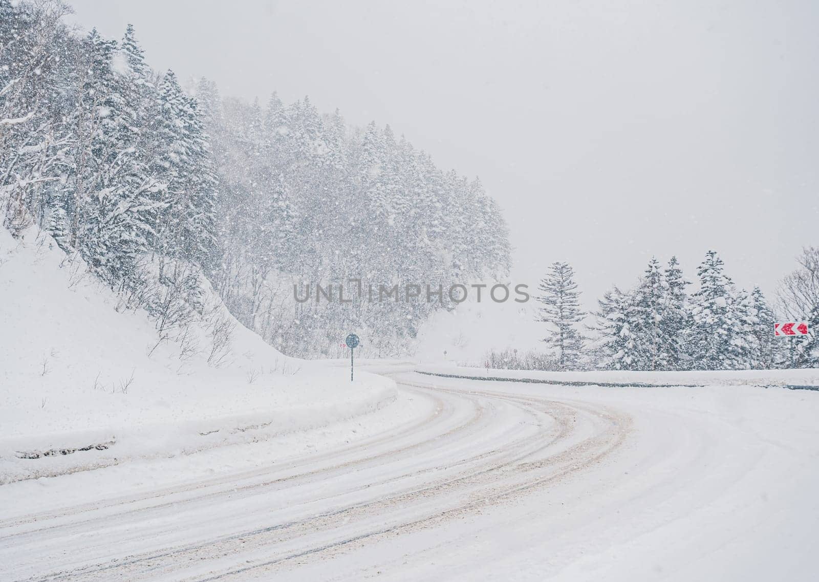 Snow is heavily falling on a winding mountain road lined with dense forest. The road appears slippery due to the accumulating snow.