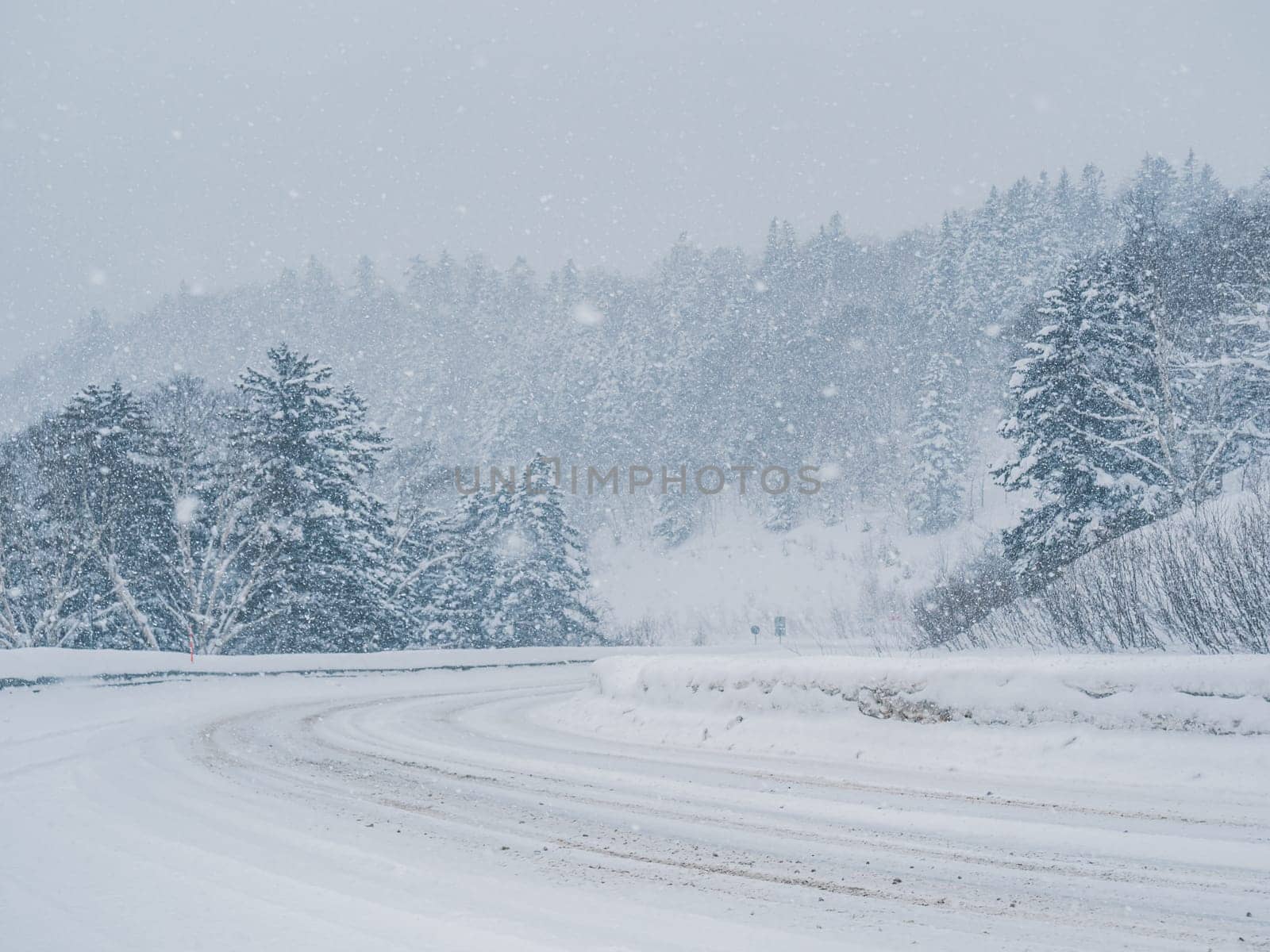 Snow is heavily falling on a winding mountain road lined with dense forest. The road appears slippery due to the accumulating snow.