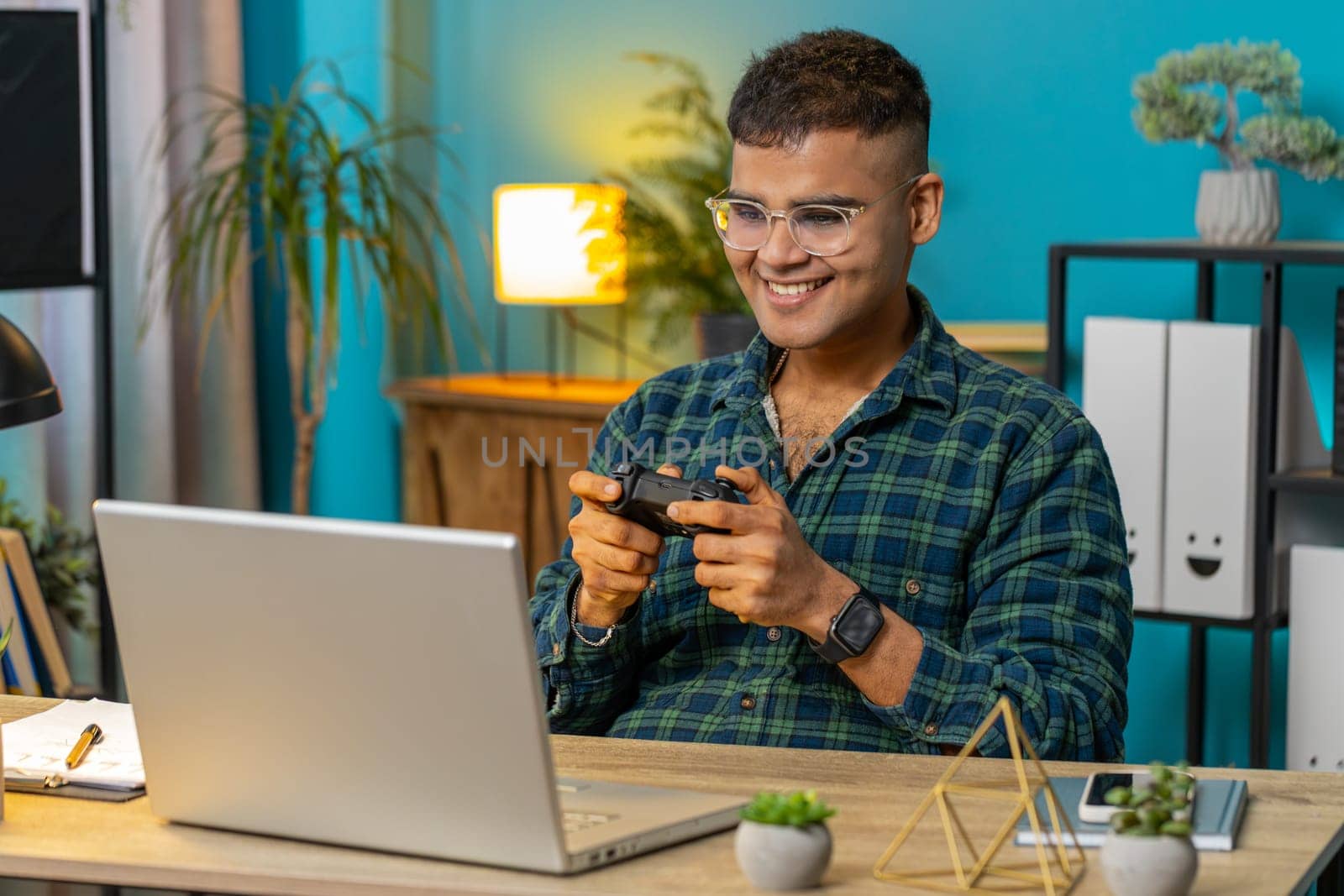 Excited Indian Hindu man playing video game on laptop spending leisure time at home office desk. Happy Hispanic freelancer guy playing video games with joystick controller on computer. Lifestyle