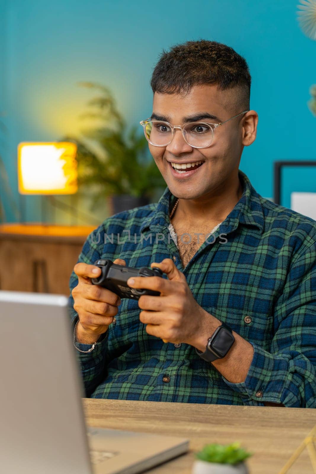 Excited Indian Arabian man playing video game on laptop spending leisure time at home office desk. Happy Hispanic freelancer guy playing video games with joystick controller on computer. Vertical