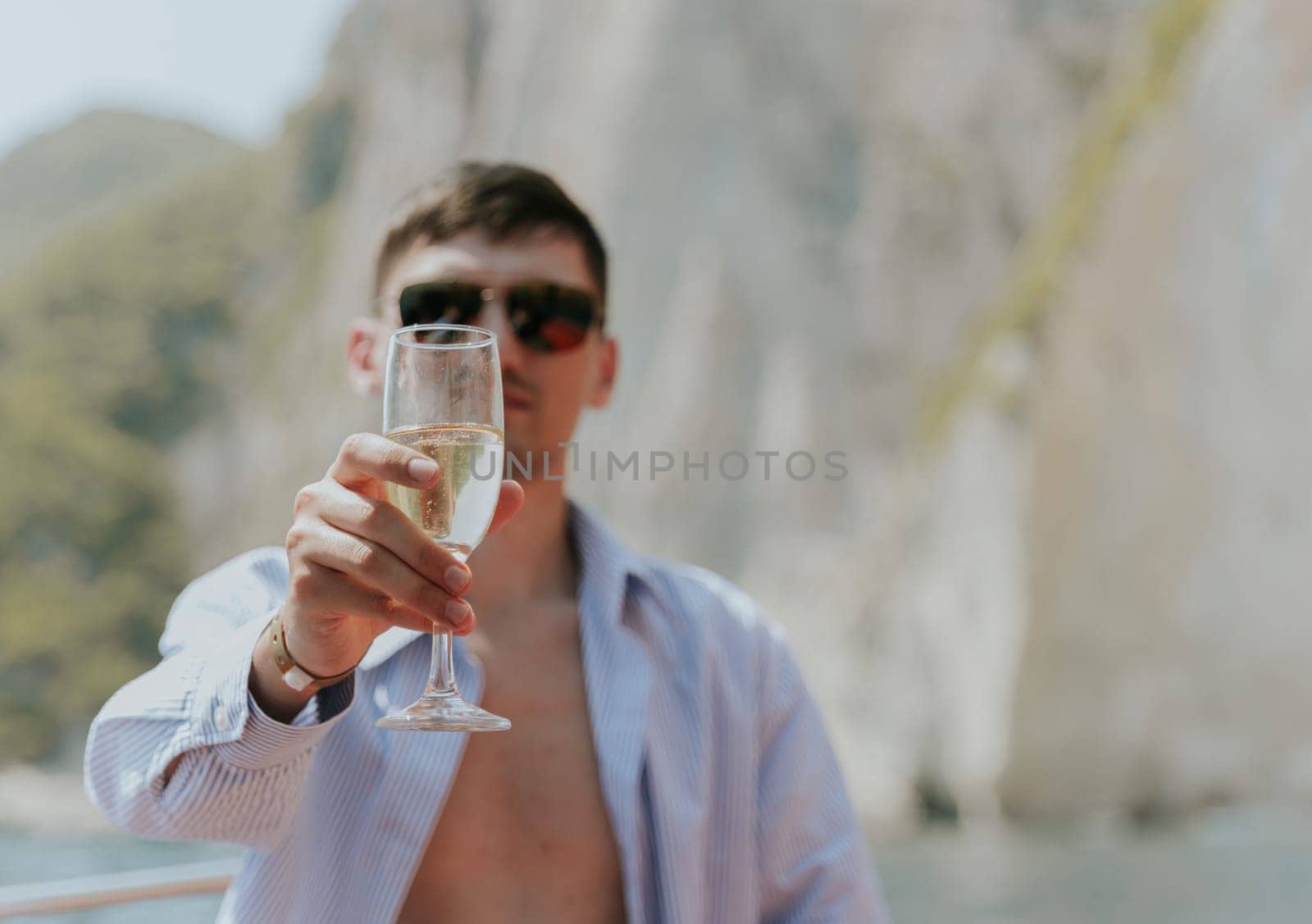 Portrait of one young Caucasian handsome guy in sunglasses sits at the stern of a boat, holds out a glass of champagne, looks into the camera and enjoys sailing on the sea on a private boat on a sunny summer day, close-up side view with selective focus.