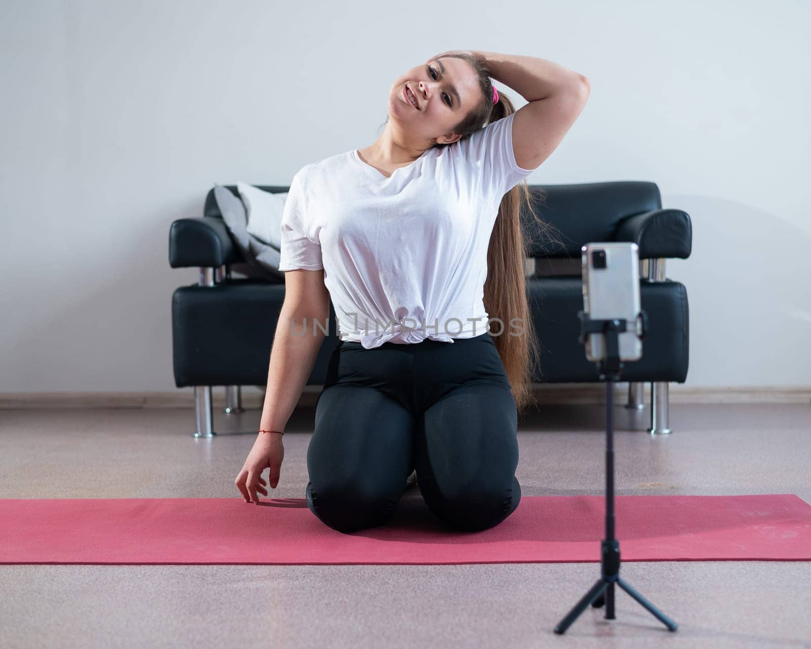 Charming plus size girl in sportswear is doing fitness exercises with an online trainer on the phone against a white background.