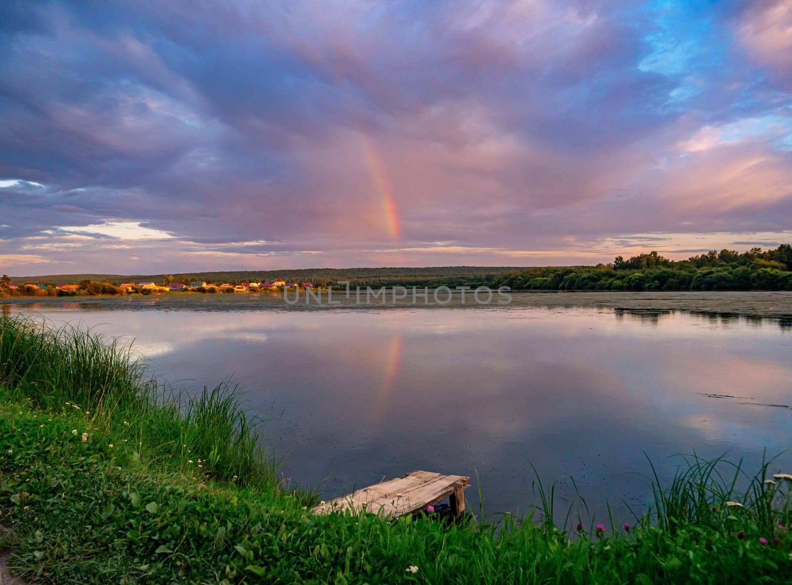 A serene countryside scene during sunset, showcasing a tranquil lake with a wooden dock in the foreground. A rainbow is reflected in the water, surrounded by a soft pink and purple sky.