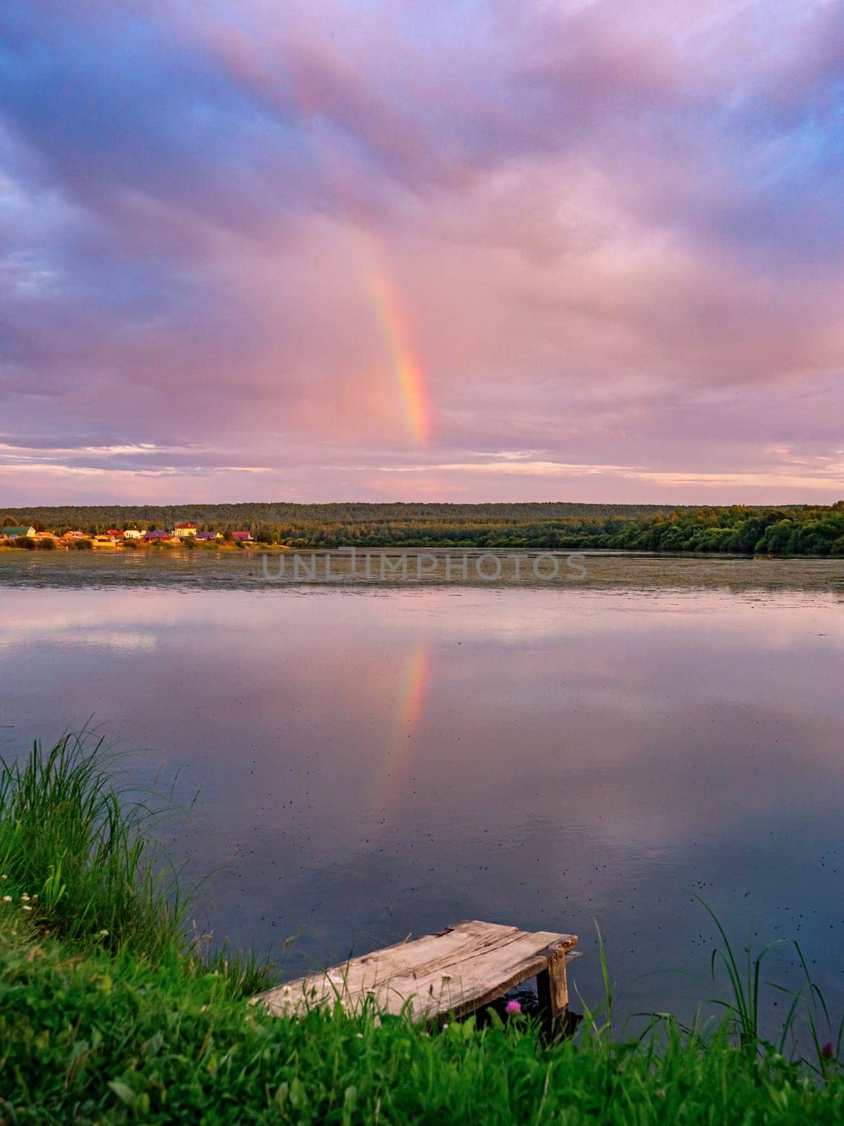 Beautiful pink sunset with rainbow reflection over calm lake at countryside evening. Tranquil lake with a wooden dock in the foreground. by Busker