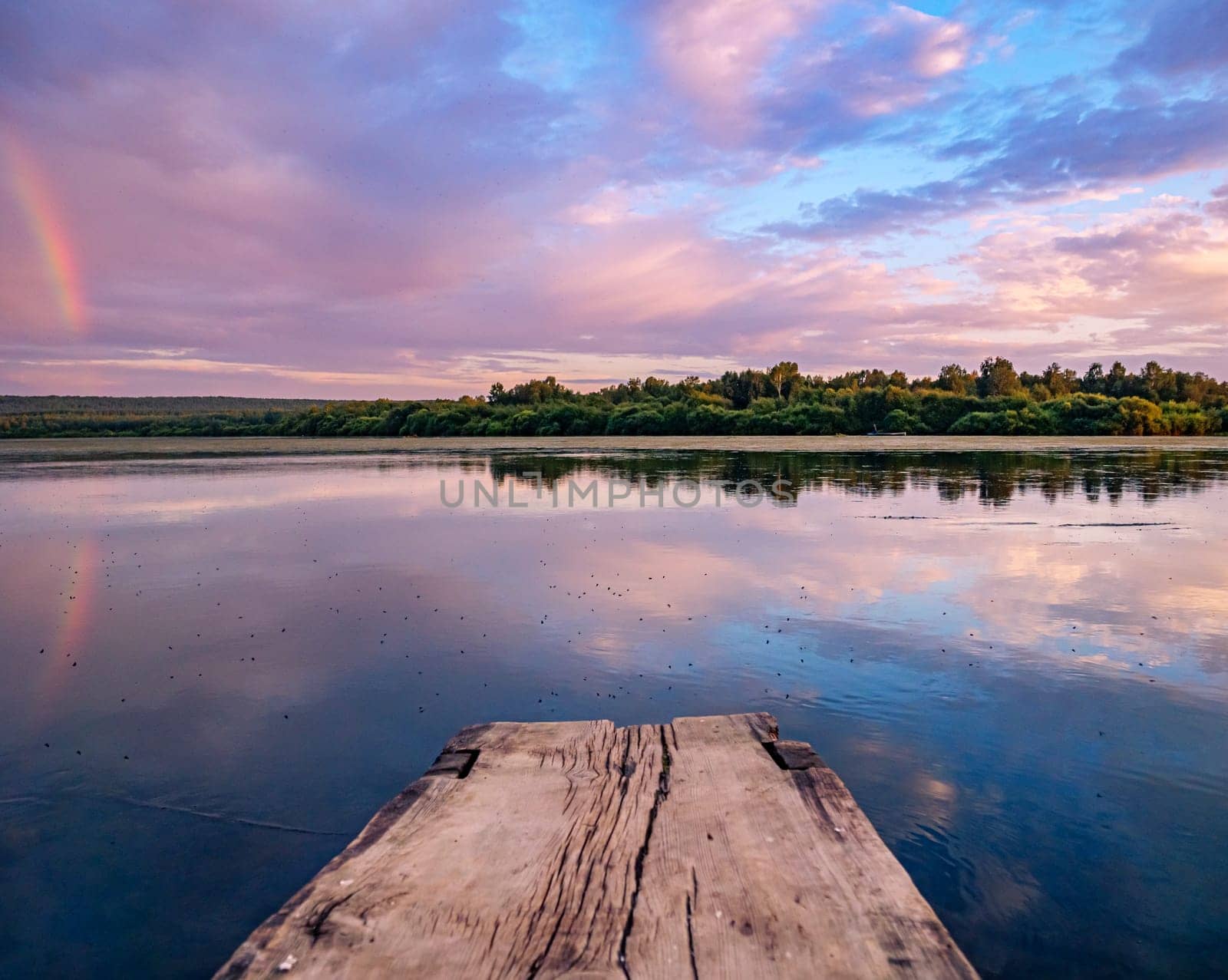 A serene countryside scene during sunset, showcasing a tranquil lake with a wooden dock in the foreground. A rainbow is reflected in the water, surrounded by a soft pink and purple sky.