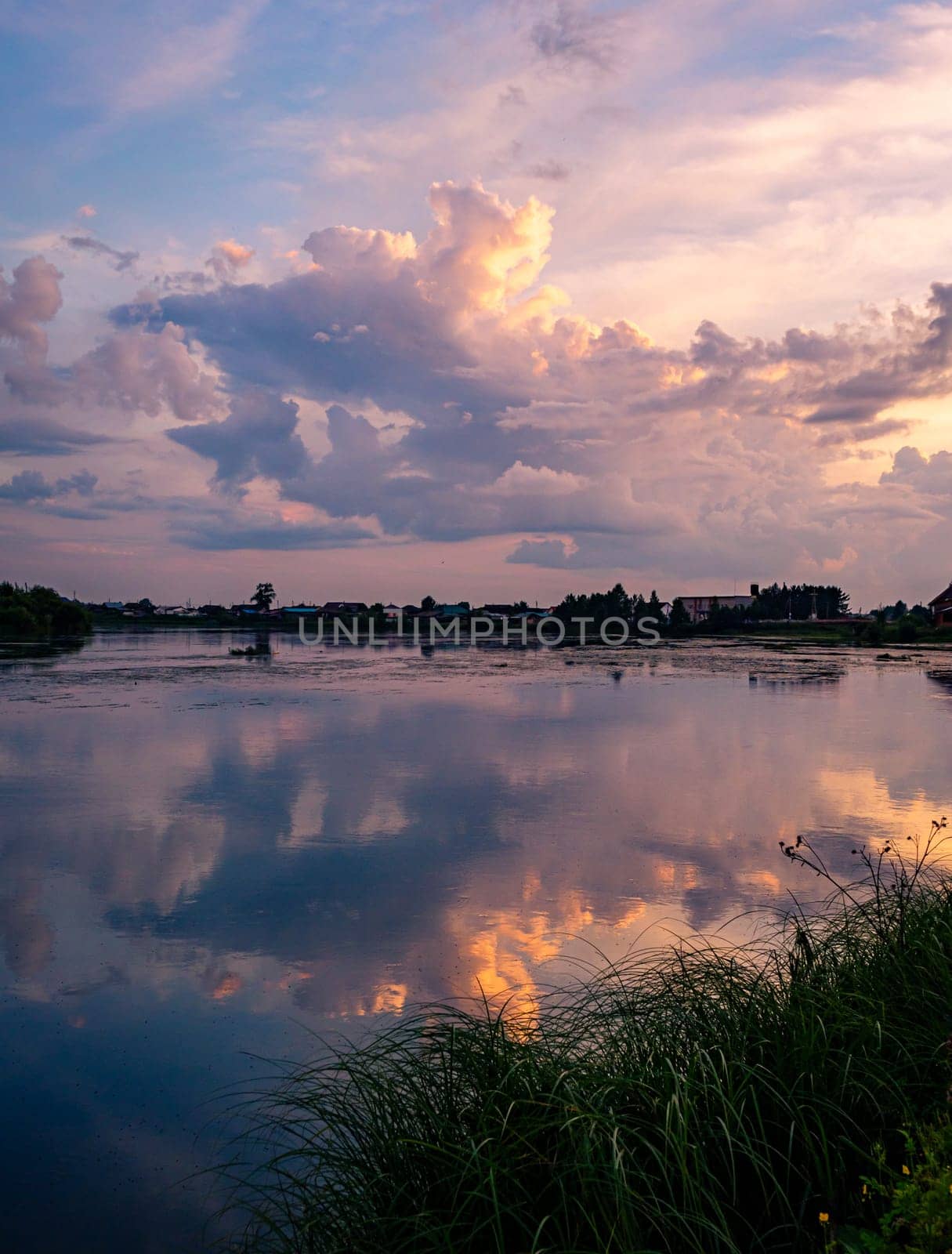 A tranquil river reflects the colorful sunset sky with clouds in a rural countryside setting. The serene scene is surrounded by lush greenery, creating a picturesque and peaceful environment.