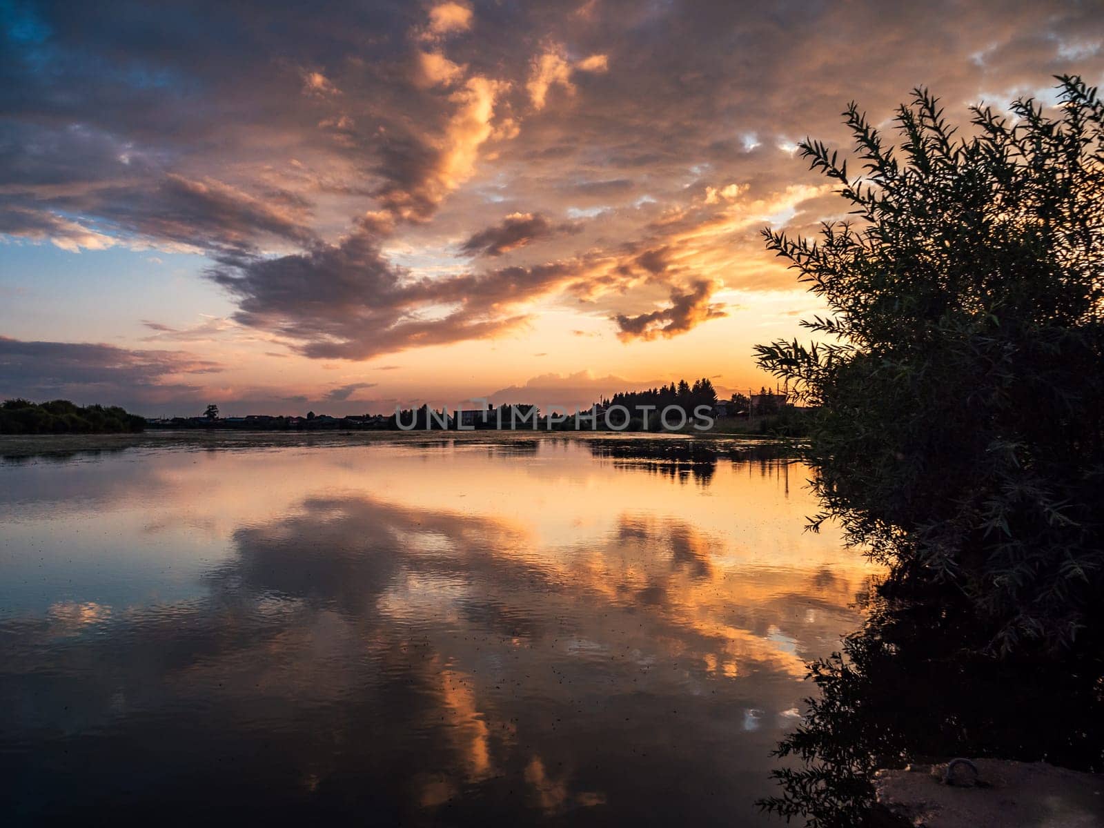 A tranquil river reflects the colorful sunset sky with clouds in a rural countryside setting. The serene scene is surrounded by lush greenery, creating a picturesque and peaceful environment.