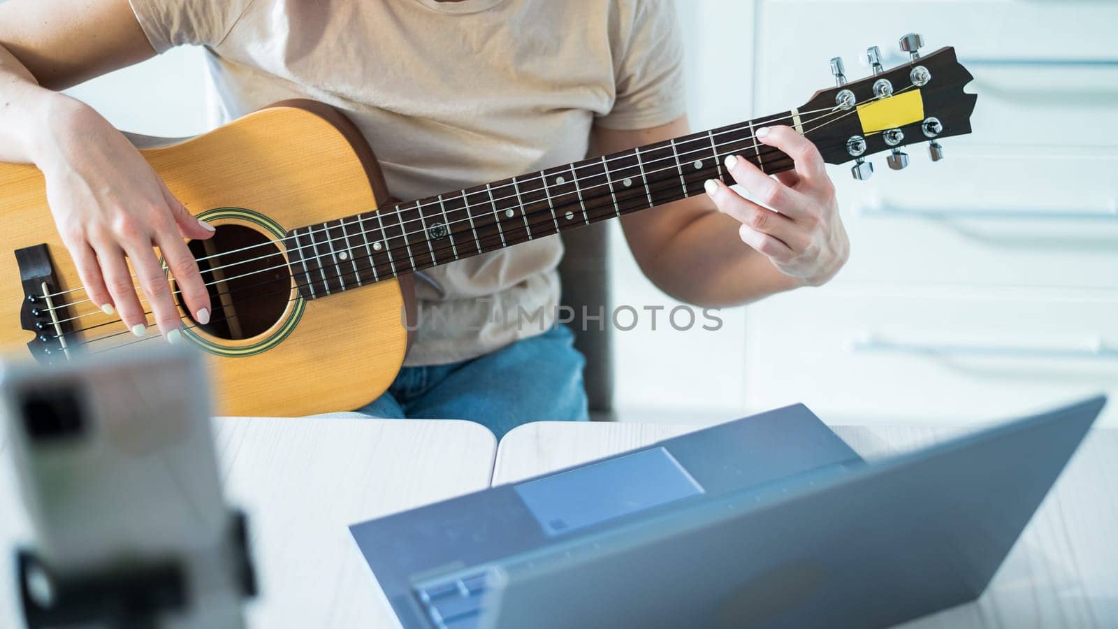 A woman sits in the kitchen during a remote acoustic guitar lesson. A girl learns to play the guitar and watches educational videos on a laptop by mrwed54
