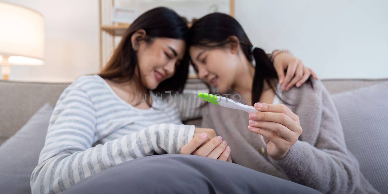 Couple smiling while looking at a positive pregnancy test on the couch at home. Women sharing a joyful moment anticipating parenthood together by nateemee