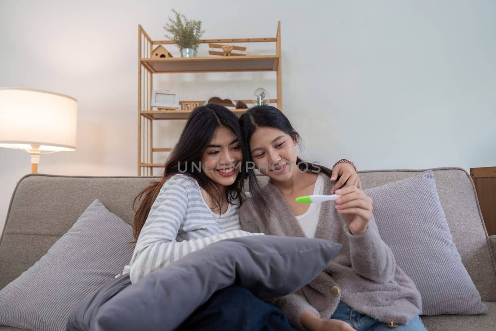 Couple smiling while looking at a positive pregnancy test on the couch at home. Women sharing a joyful moment anticipating parenthood together.