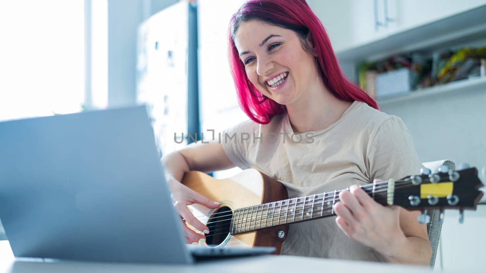 A woman sits in the kitchen during a remote acoustic guitar lesson. A girl learns to play the guitar and watches educational videos on a laptop by mrwed54