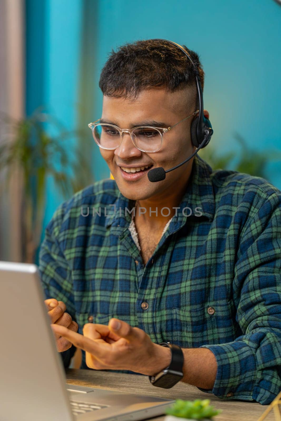 Indian business man wearing headset freelance worker call center or support service operator helpline talking with client or colleague communication support. Guy sitting at home office desk. Vertical