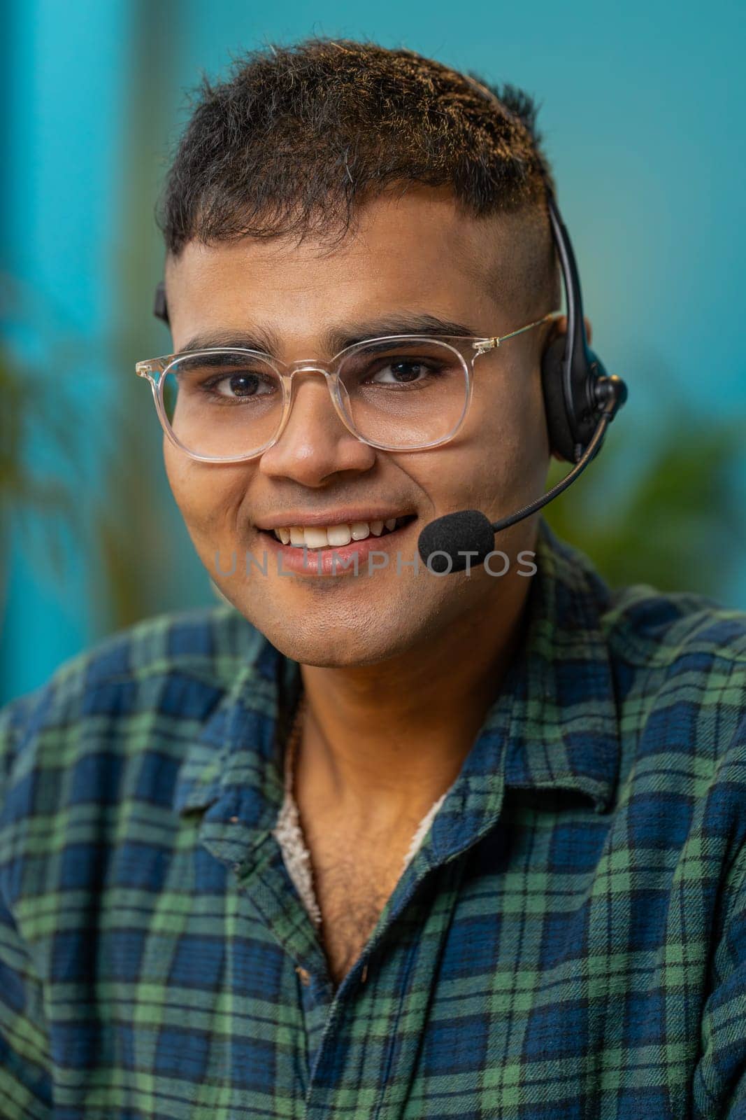 Indian business man wearing headset freelance worker call center or support service operator helpline talking with client or colleague communication support. Guy sitting at home office desk. Vertical