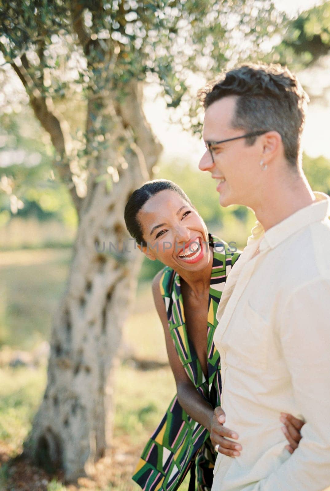 Laughing woman looking at man hugging his waist near green tree in garden. High quality photo