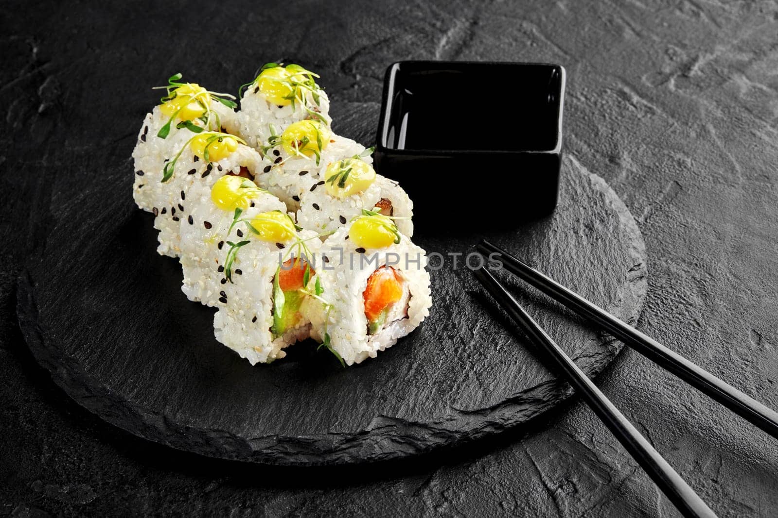 Sesame coated sushi rolls with avocado and fresh salmon topped with drop of mayo and microgreens traditionally served with soy sauce on black slate board. Japanese cuisine