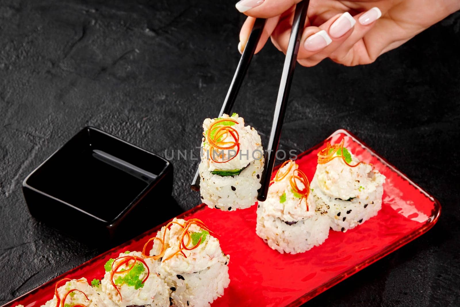 Female hand gracefully holding sushi roll with creamy crab topping with chopsticks over vibrant red serving plate on dark textured background, ready to dip in soy sauce and enjoy Japanese style snack