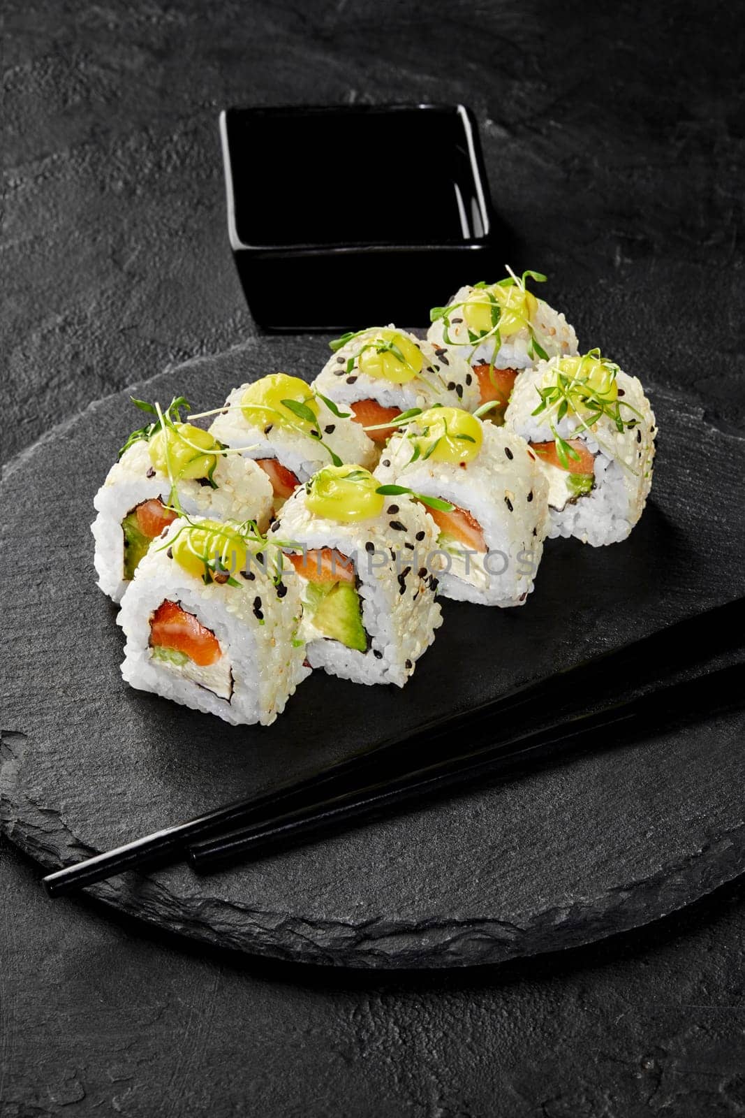 Colorful sesame coated sushi rolls filled with salmon, avocado and cream cheese garnished with spicy mayo and greens traditionally served with soy sauce on black slate serving board. Japanese cuisine