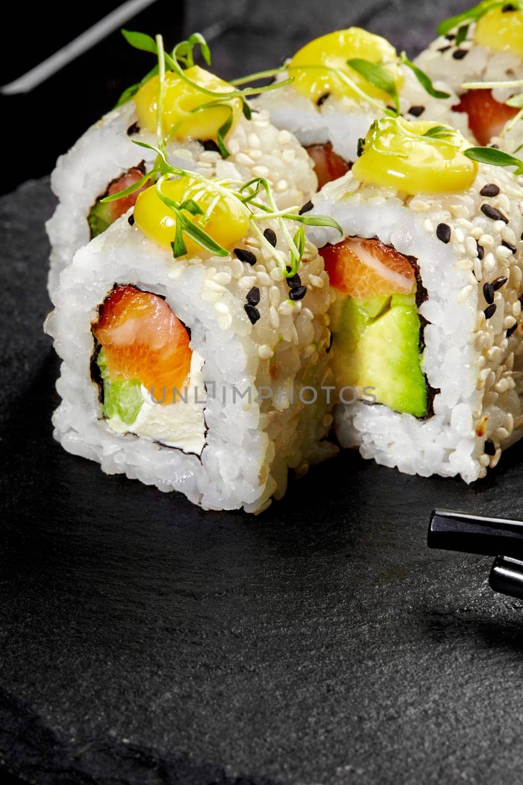 Closeup of classic sesame coated uramaki rolls filled with cream cheese, fresh salmon and avocado, topped with spicy mayo drops and microgreens, served on black stone board