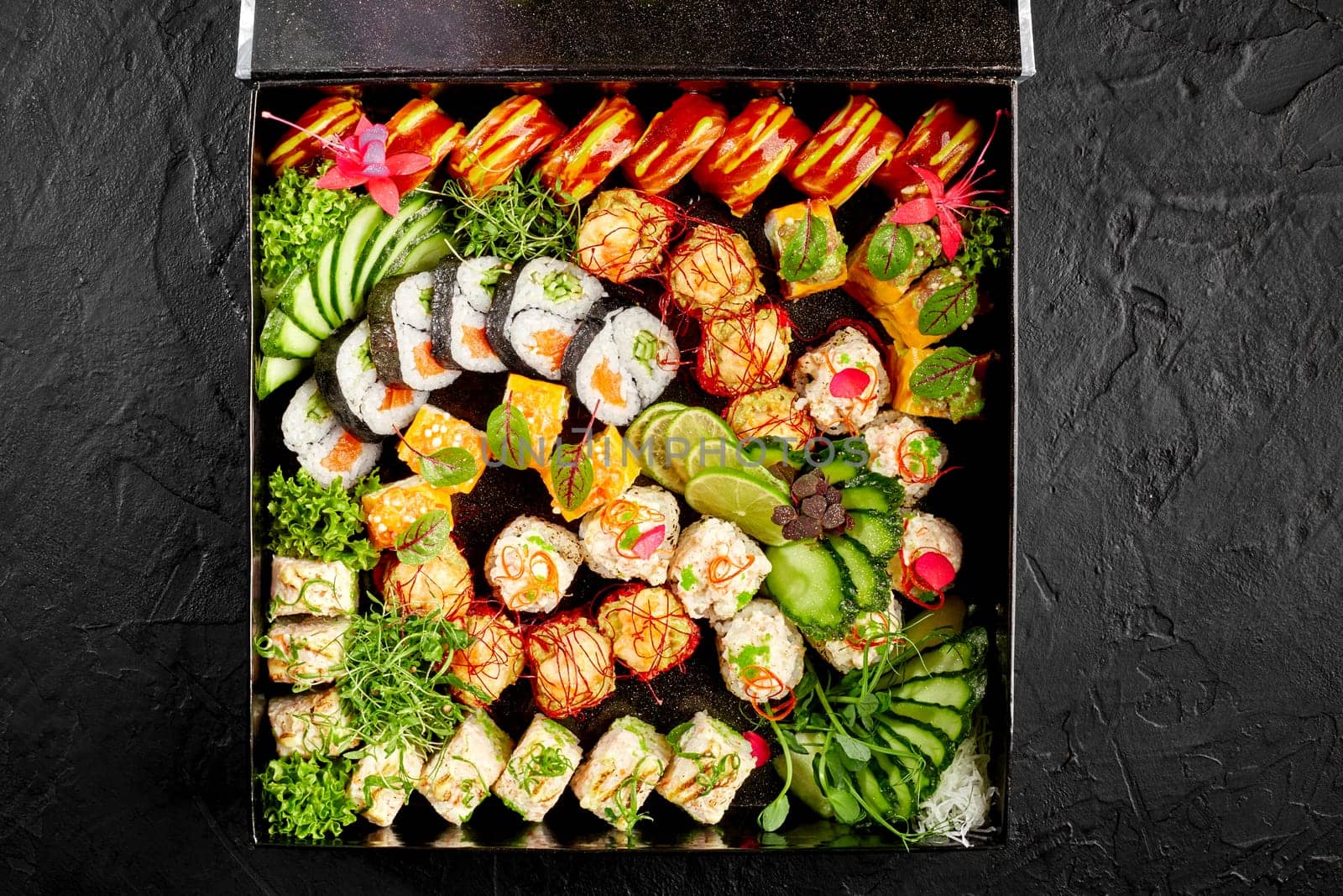 Delightful sushi set featuring array of rolls, embellished with fresh garnishes, vegetables and vibrant flowers, arranged in box against black textured background. Delivery or takeaway food concept