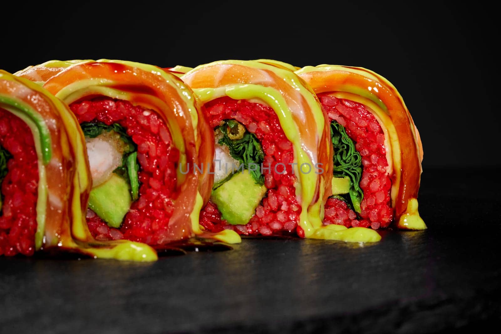 Closeup of vibrant red rice sushi rolls with tempura shrimp and avocado, topped with salmon slice and spicy sauces, presented on black slate board. Japanese fusion cuisine