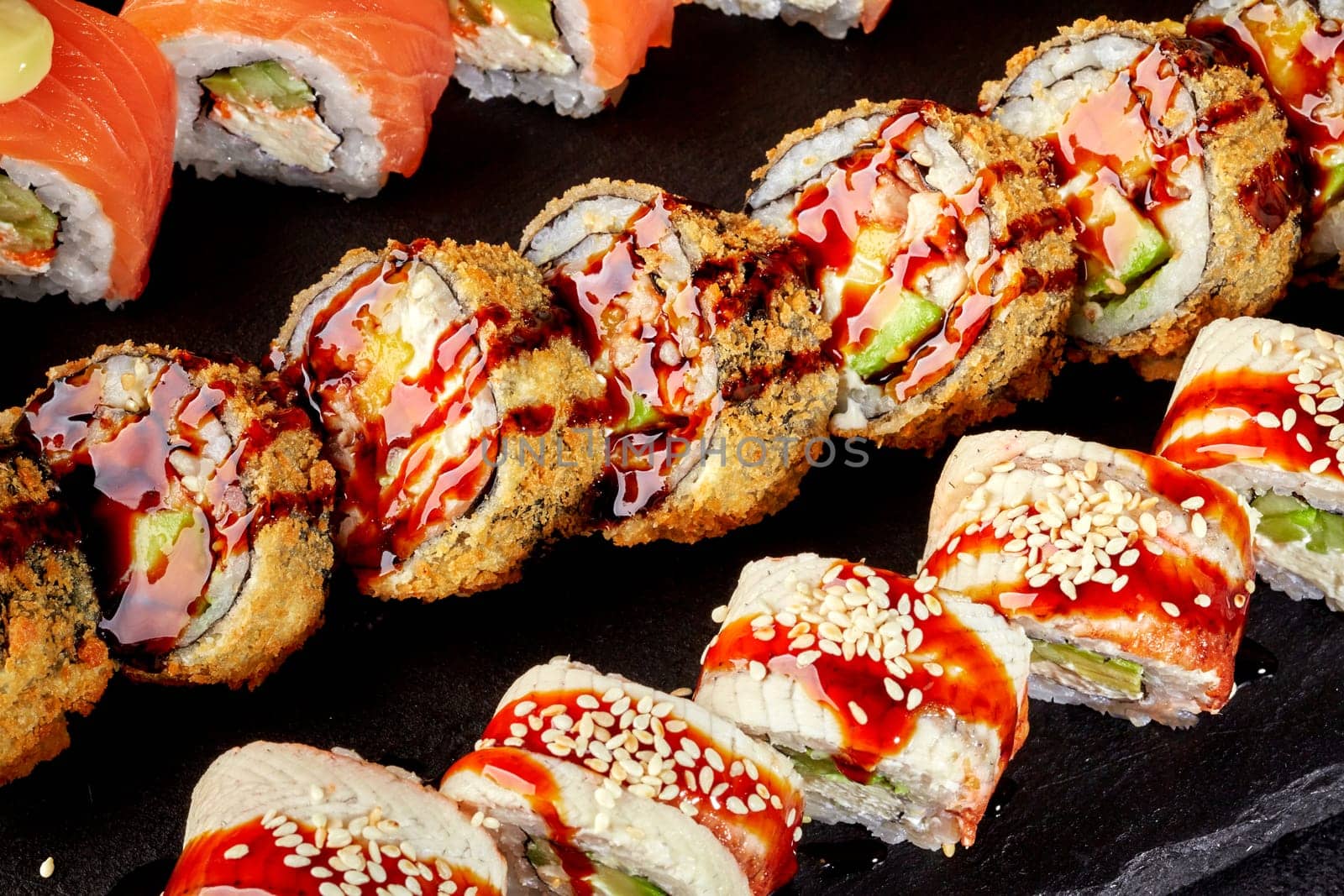Appetizing crispy tempura rolls drizzled with unagi sauce, accompanied by ell and salmon topped uramaki served on black slate board, closeup detail view. Popular Japanese style snack