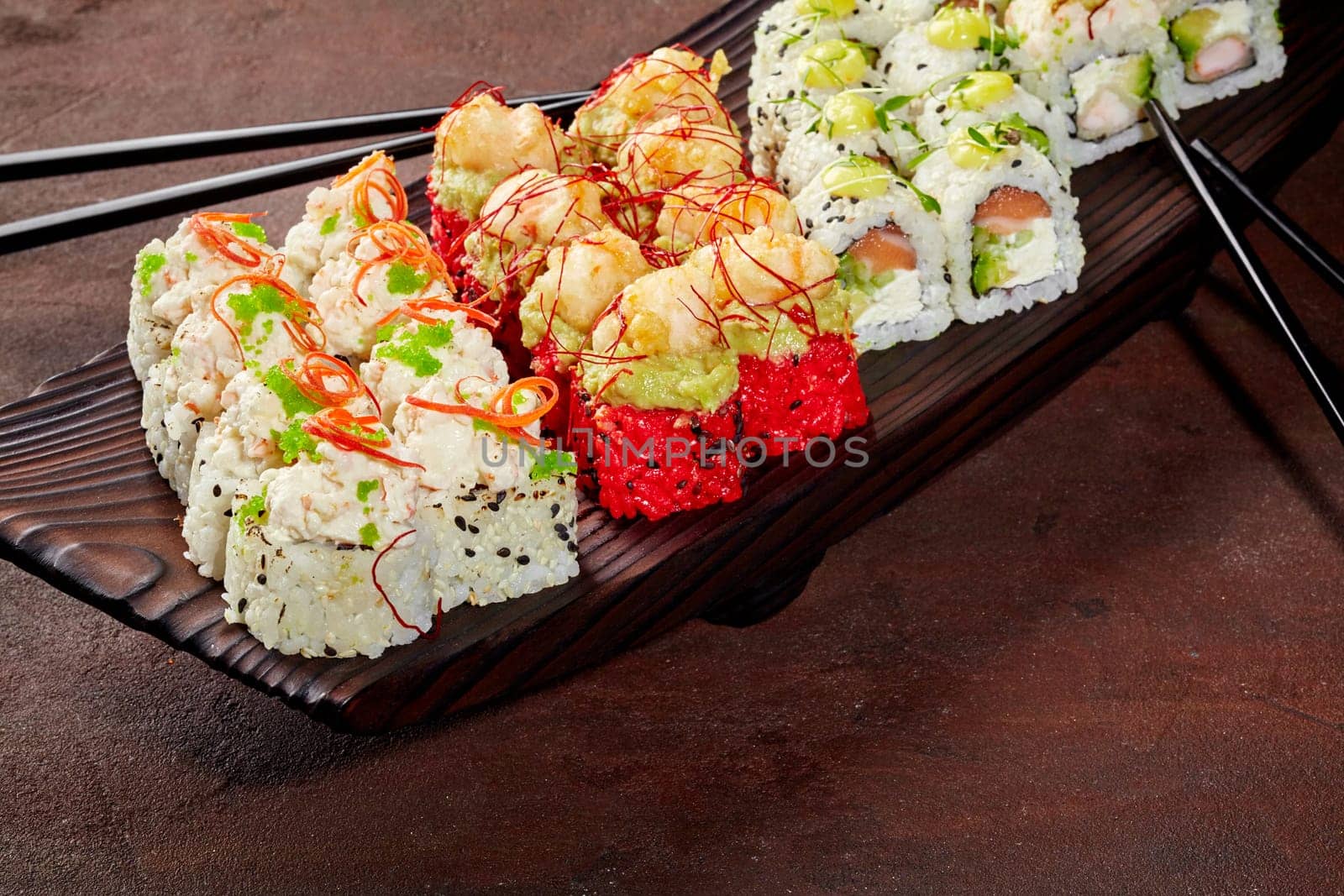 Enticing set of sushi rolls with salmon, shrimp, tobiko and sesame garnished with greens and spicy sauce, arranged on dark wooden serving tray. Stylish presentation of popular Japanese dish