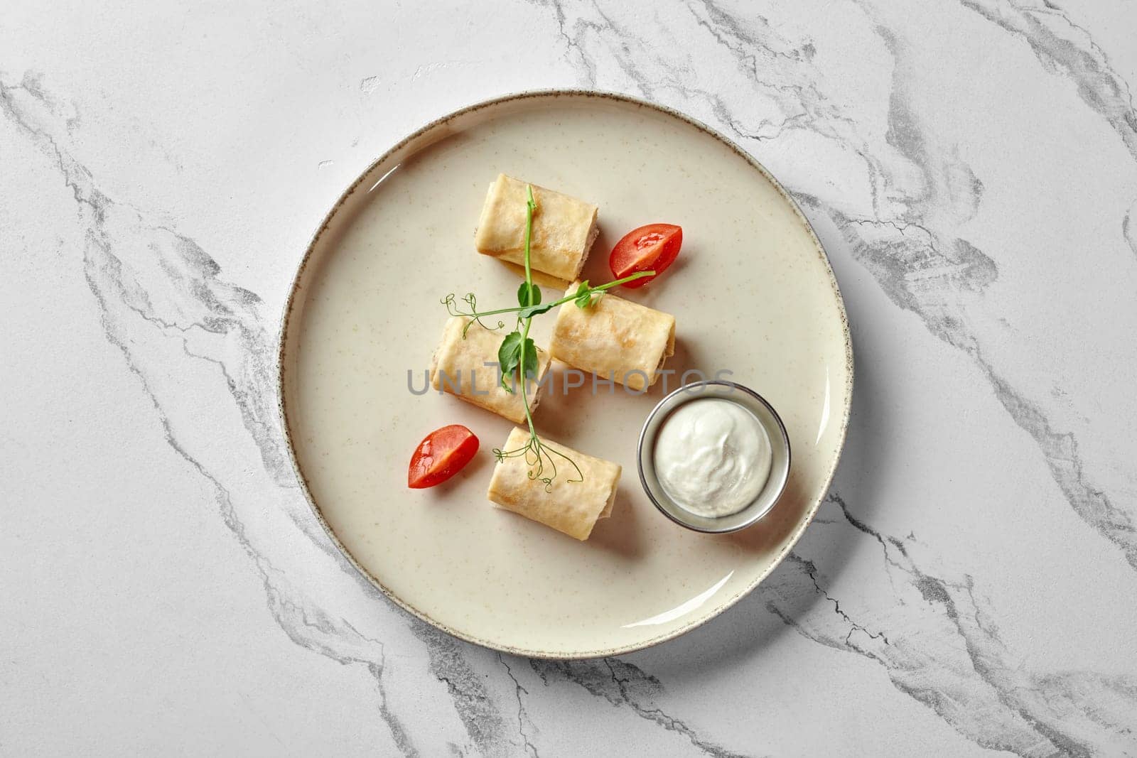 Top view of savory chicken stuffed crepe rolls garnished with fresh herbs and cherry tomato, served with creamy dipping sauce on ceramic plate against marble background