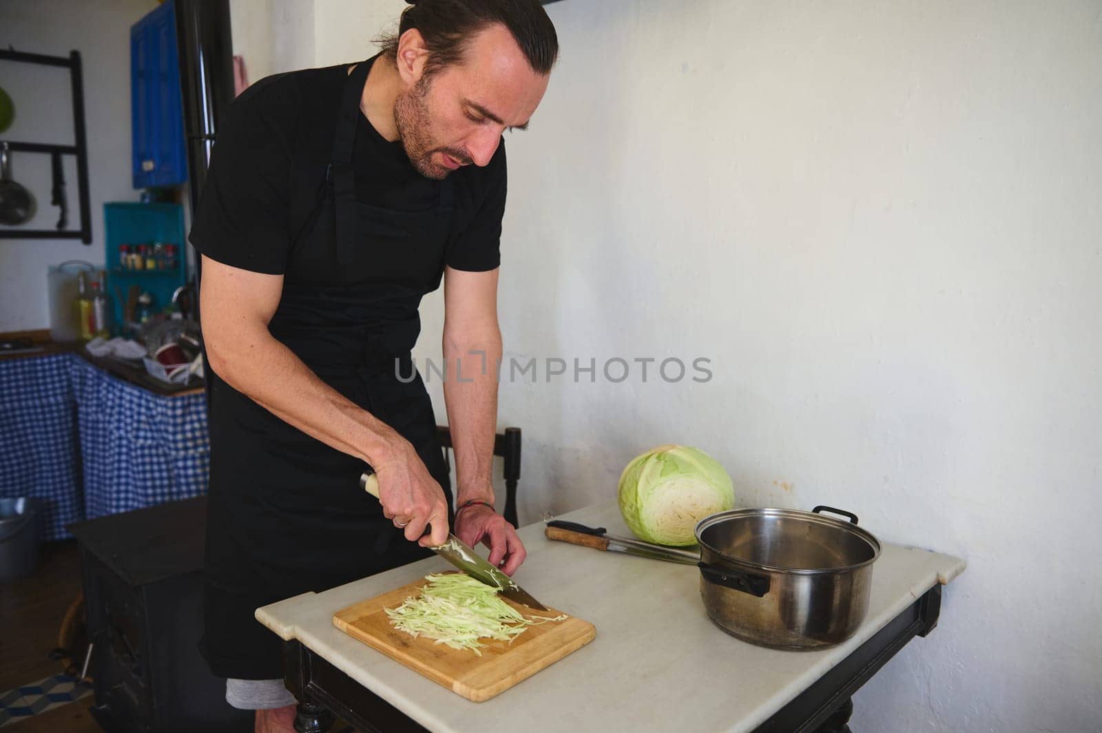 Handsome Caucasian bearded male chef chopping raw fresh cabbage on a wooden board in rustic home kitchen. Food concept