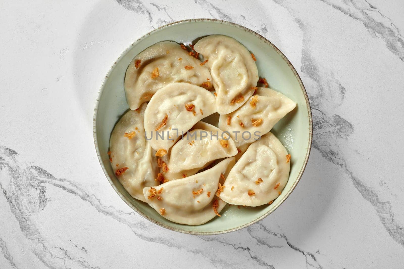 Traditional Ukrainian vareniki stuffed with ground meat garnished with crispy fried onions served in rustic ceramic bowl against marble background. Authentic cuisine