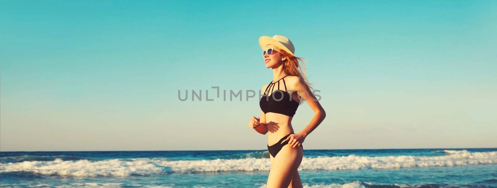 Summer vacation, happy smiling woman walking running in bikini swimsuit and straw hat on the beach on sea coast with waves background on sunny day, blue sky