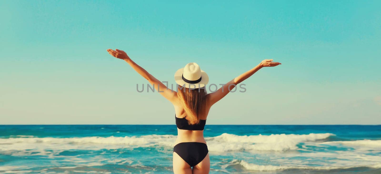 Summer vacation, rear view of beautiful happy slim woman in bikini swimsuit and straw hat raising her hands up on the beach on sunny day on sea coast background