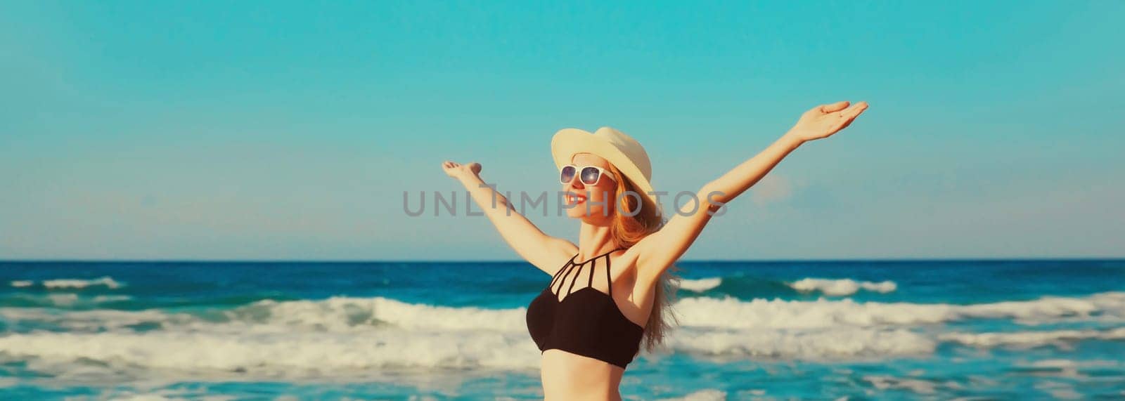 Summer vacation, happy smiling woman in bikini swimsuit and straw hat raising her hands up on the beach on sea coast background