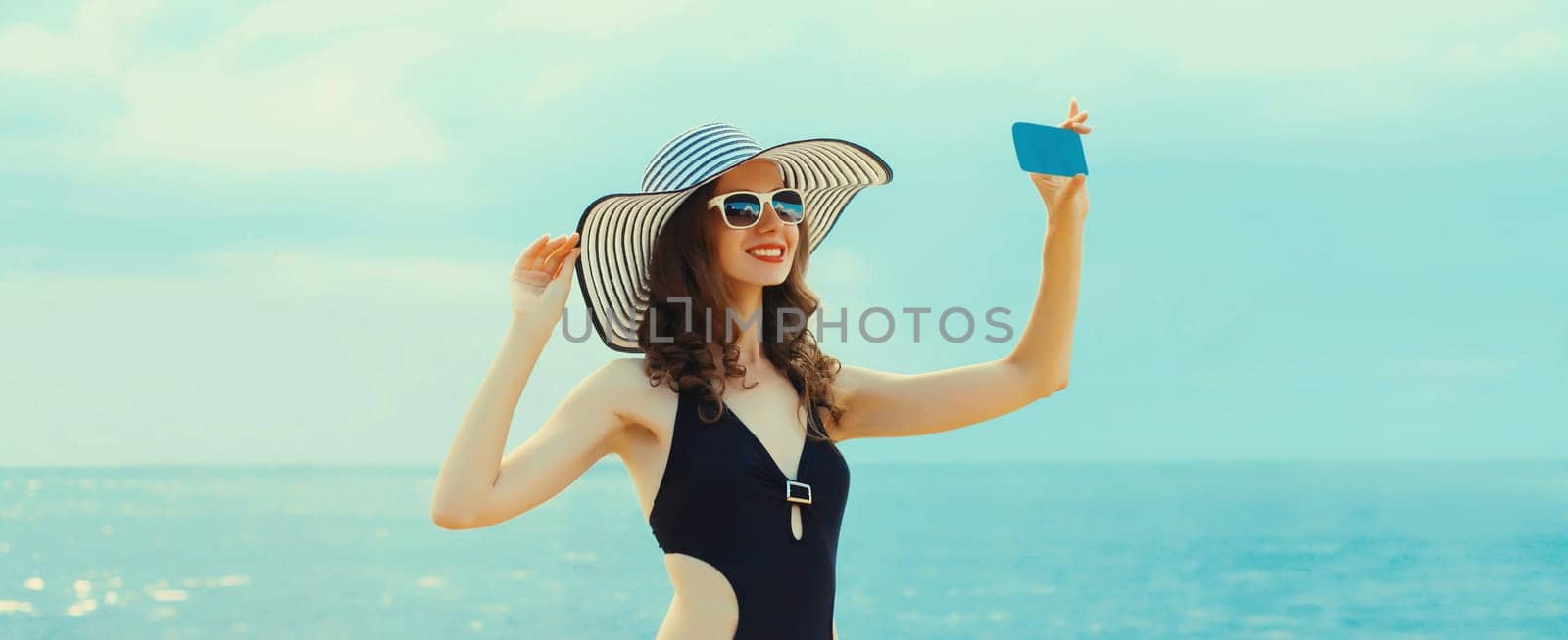 Summer portrait of beautiful young woman taking selfie with smartphone on the beach wearing straw hat and bikini on sea background