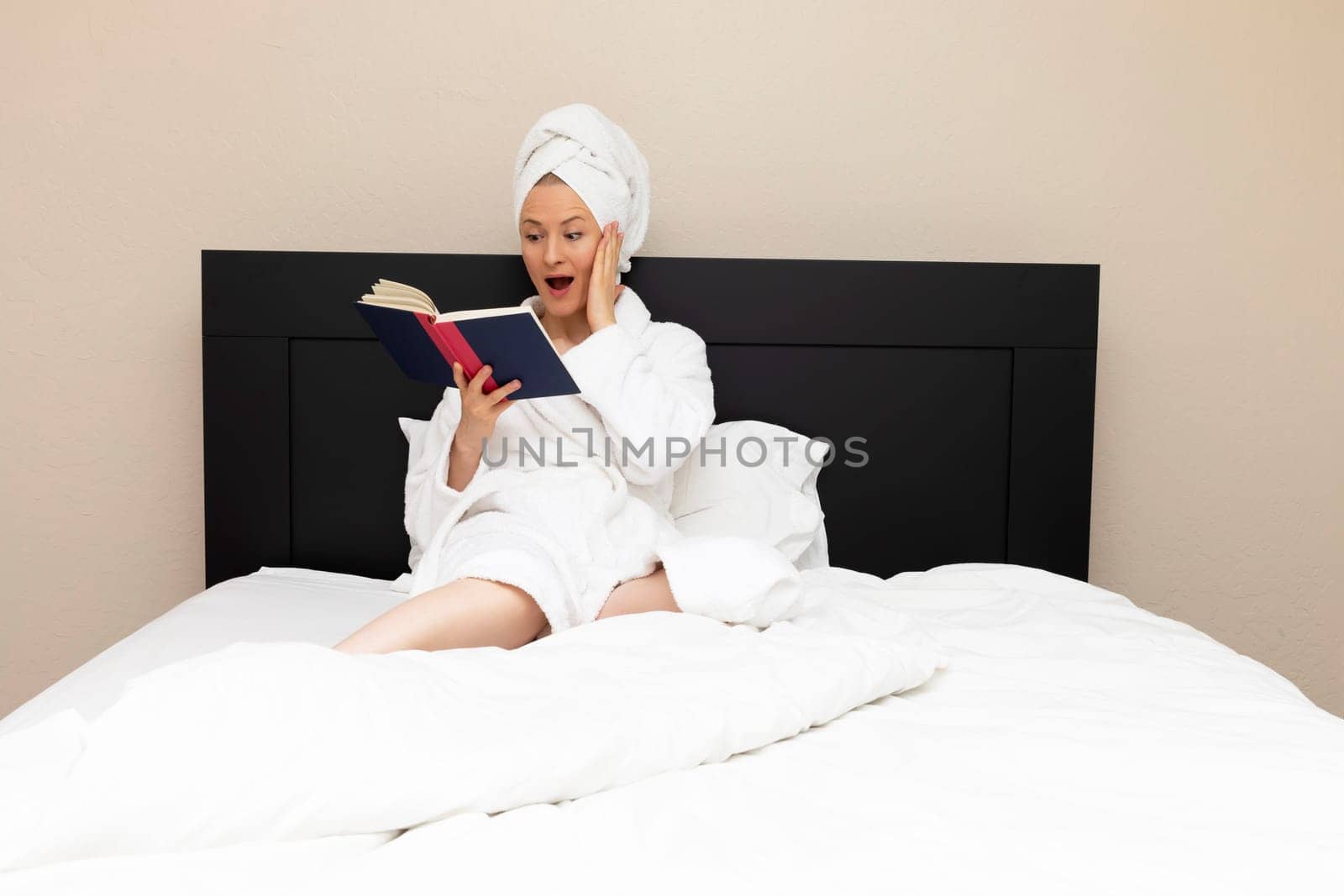 Adult Woman Sits In Bed with Surprised, Shocked Facial Expression while Reading A Book. Emotional Expression Of Face, Caucasian Mature Female Reading Horror Novel Fiction, Detective Story. Book Lover
