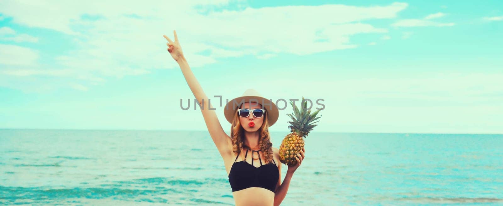 Summer vacation, beautiful happy young woman in bikini swimsuit and straw hat holding pineapple fruits blowing kiss on the beach on sea coast background