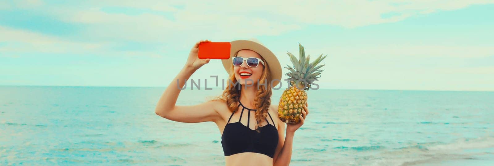 Summer vacation, happy smiling young woman holding pineapple taking selfie with smartphone wearing bikini swimsuit and straw hat on the beach on sea coast background