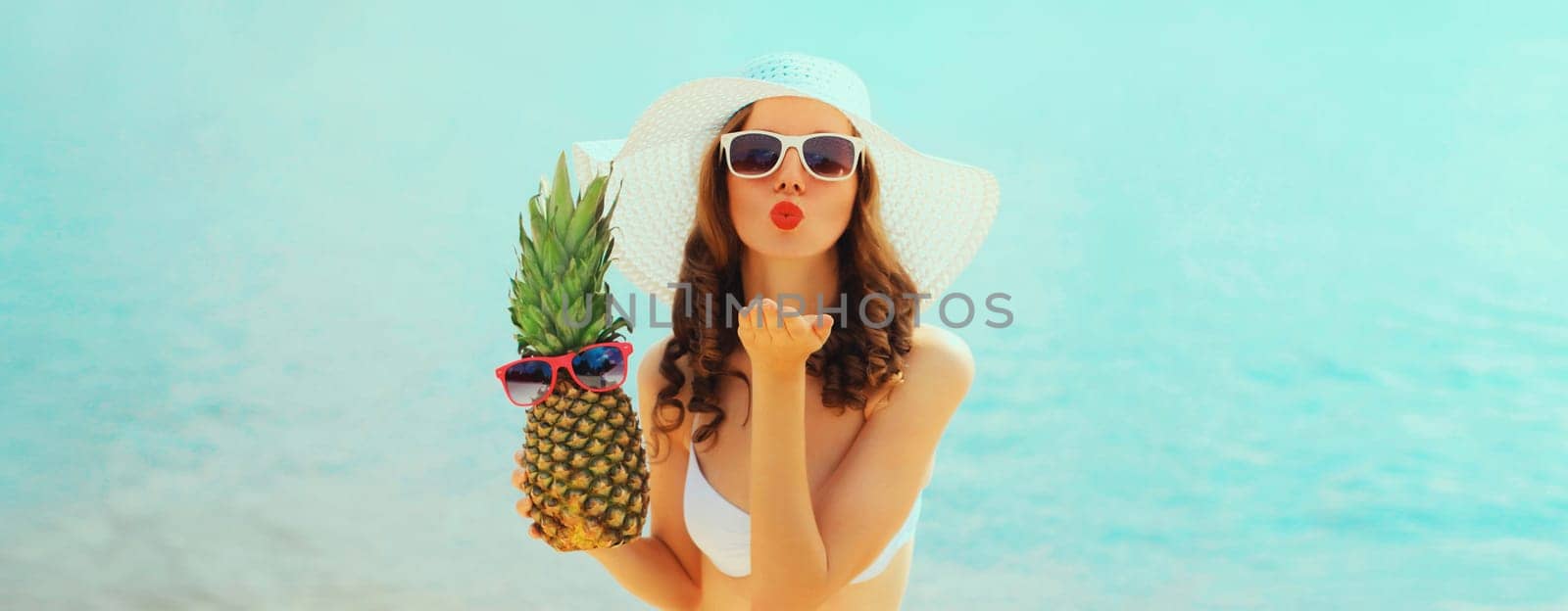 Summer vacation, beautiful happy young woman in bikini swimsuit and straw hat holding pineapple fruits blowing her lips sends kiss on the beach on sea coast background on sunny day