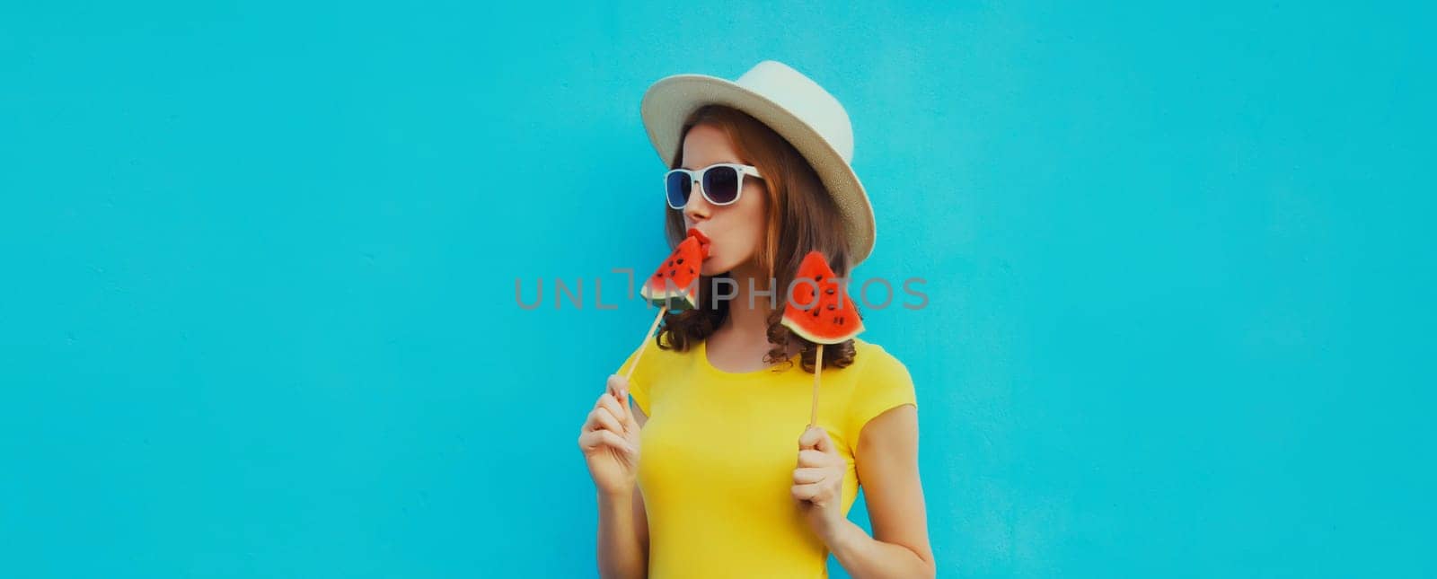 Summer portrait of stylish young woman with sweet juicy lollipop or ice cream shaped slice of watermelon wearing straw hat on blue studio background