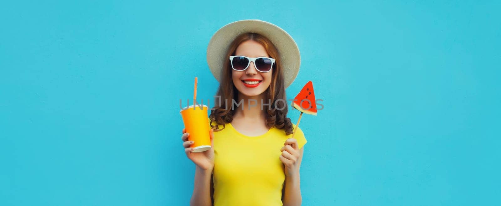 Summer portrait of happy smiling woman with cup of juice and sweet lollipop or ice cream shaped slice of watermelon in straw hat on blue background