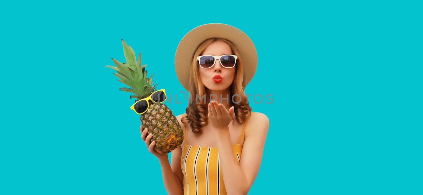 Summer portrait of beautiful young woman blowing a kiss with pineapple wearing straw hat, glasses posing on blue studio background