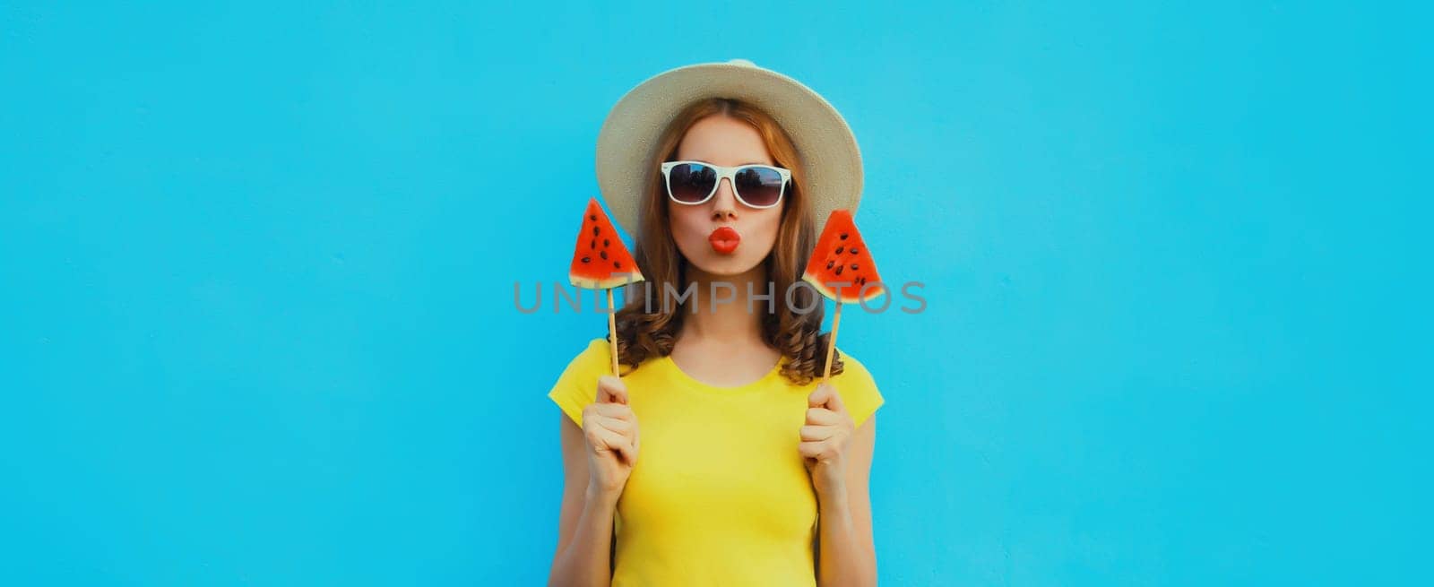 Summer portrait of stylish young woman with sweet juicy lollipop or ice cream shaped slice of watermelon wearing straw hat on blue studio background