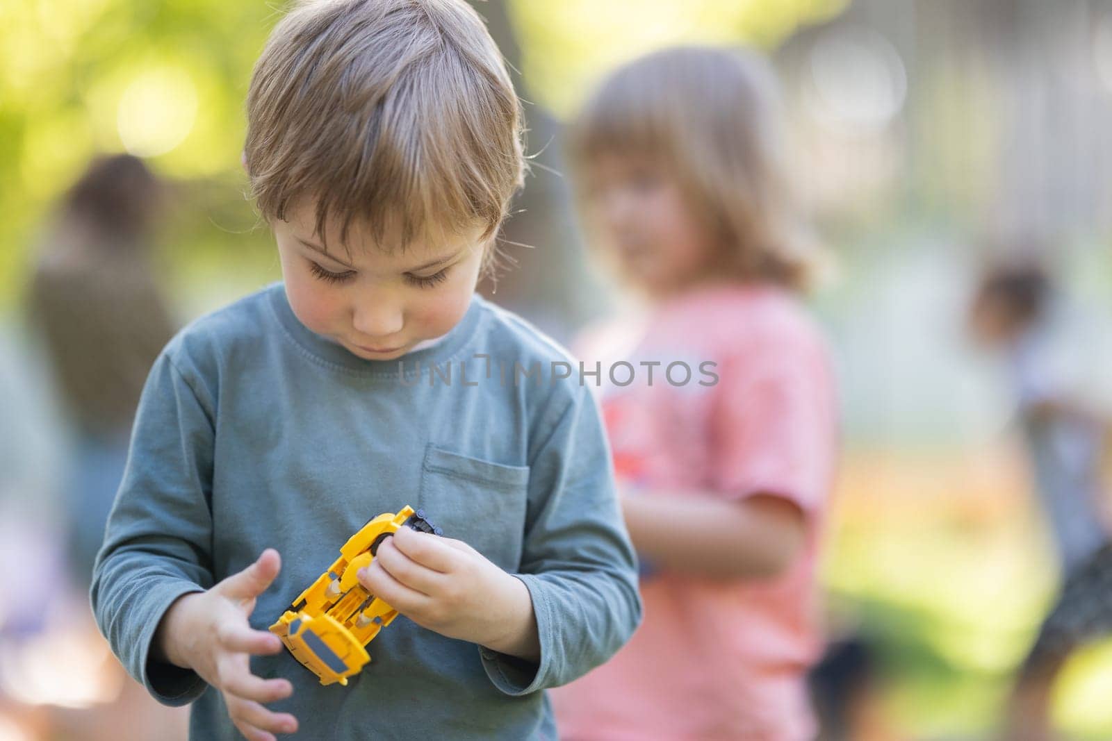 A boy is holding a toy car in his hand. He is looking at the camera. There are other children in the background