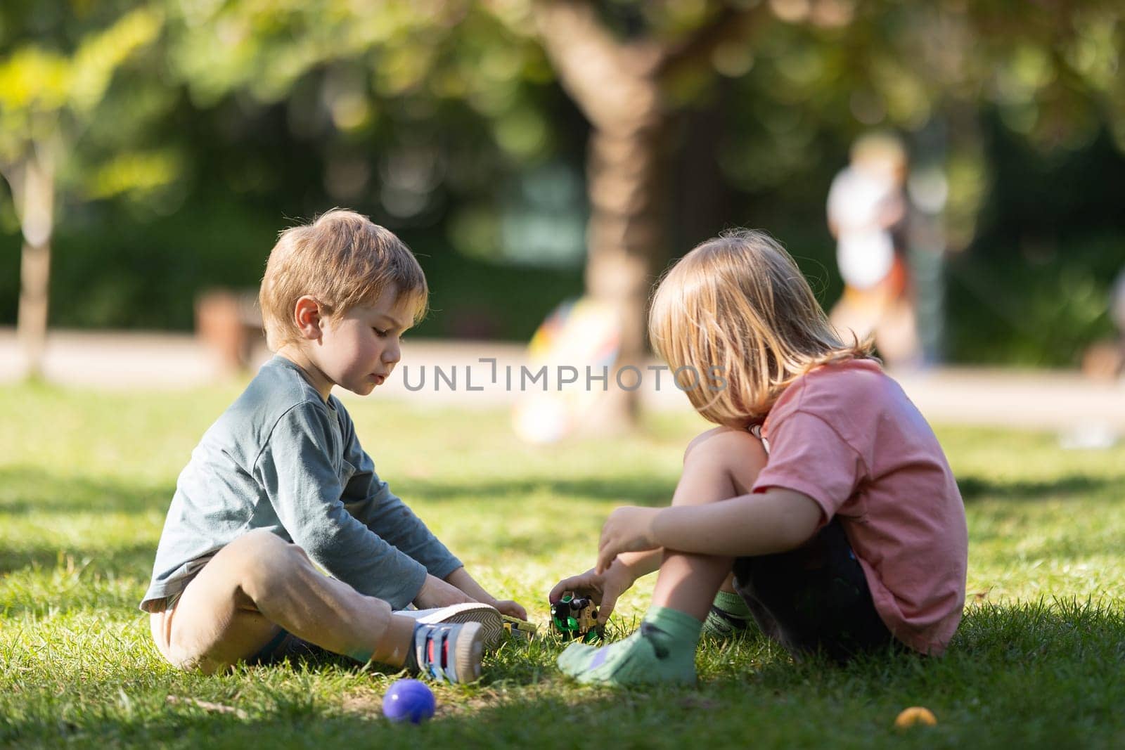 Two children are playing in a park, one of them is holding a green ball by Studia72