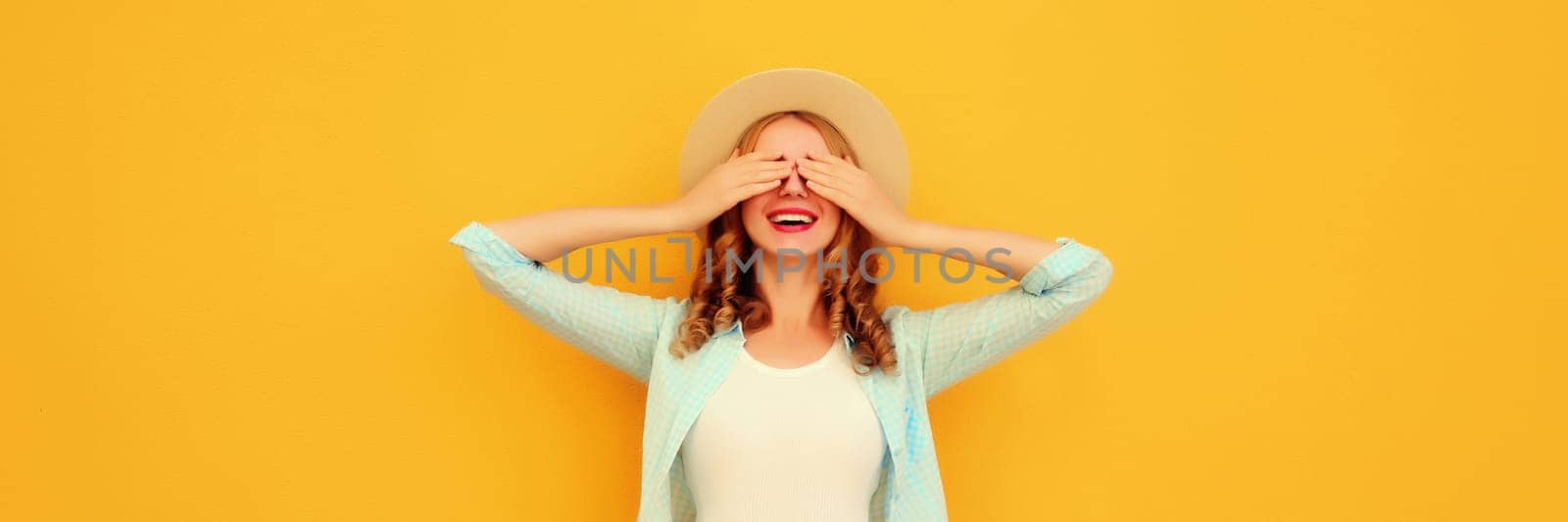 Stylish surprised young woman covering her eyes with hands wearing summer straw hat, jean jacket posing on yellow background