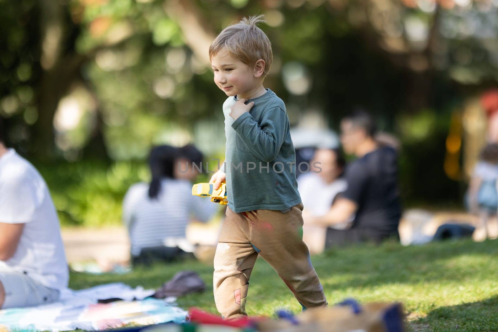 A young boy is playing with a toy car in a park. There are other people in the background, some of whom are sitting on blankets. The scene is lively and playful