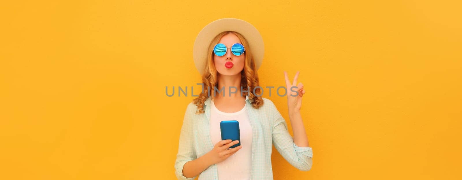 Portrait of stylish young woman posing blowing a kiss with smartphone wearing summer straw hat posing on yellow background